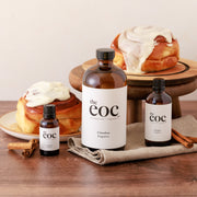 Bottles of 'the eoc' cinnamon fragrance with cinnamon rolls on a wooden surface.