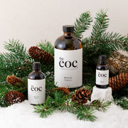 Three bottles of 'the eoc' with labels surrounded by pine branches and pinecones on a snowy background.
