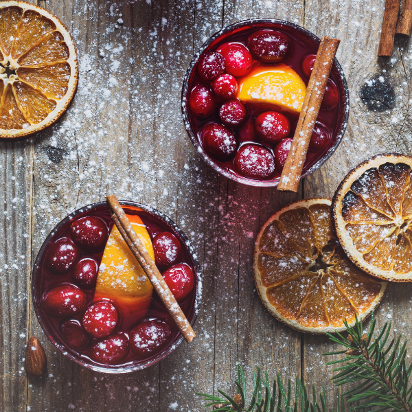 Two bowls of festive fruit punch with cranberries, oranges, and cinnamon sticks on a wooden surface with dried oranges and pine branches.