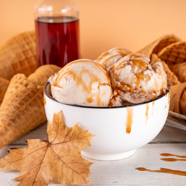 Bowl of ice cream with maple caramel drizzle surrounded by waffle cones and a glass of syrup on a wooden surface.